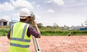 Construction worker in a hi-vis vest and white helmet uses a survey instrument on a dirt construction site, with another worker in the background.